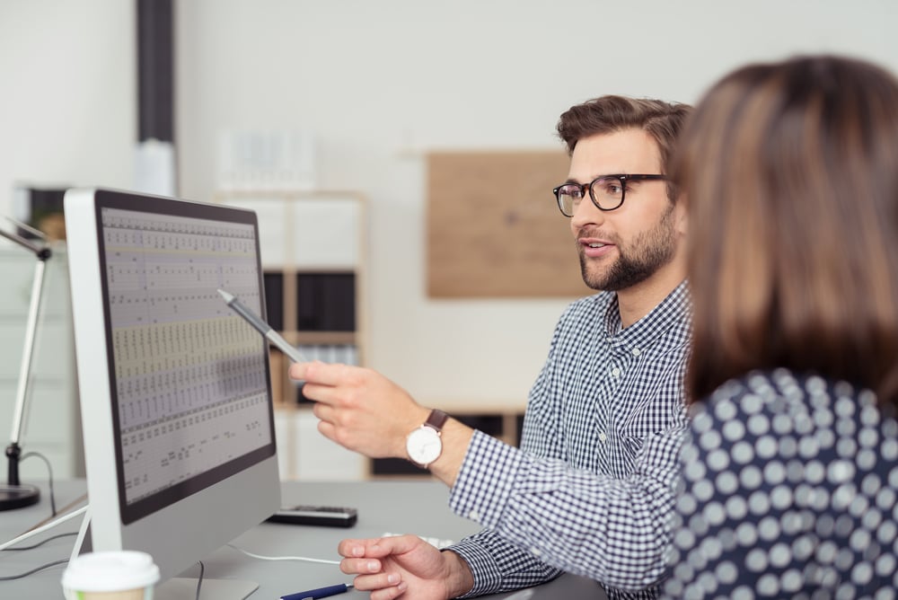 Proficient young male employee with eyeglasses and checkered shirt, explaining a business analysis displayed on the monitor of a desktop PC to his female colleague, in the interior of a modern office-3