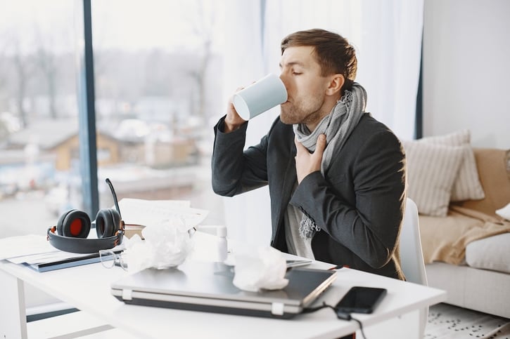  Mitarbeiter mit Grippe sitzt müde an ihrem Schreibtisch im Büro auf der vorderen Seite des Bildes, umgeben von Taschentüchern und Medikamenten.